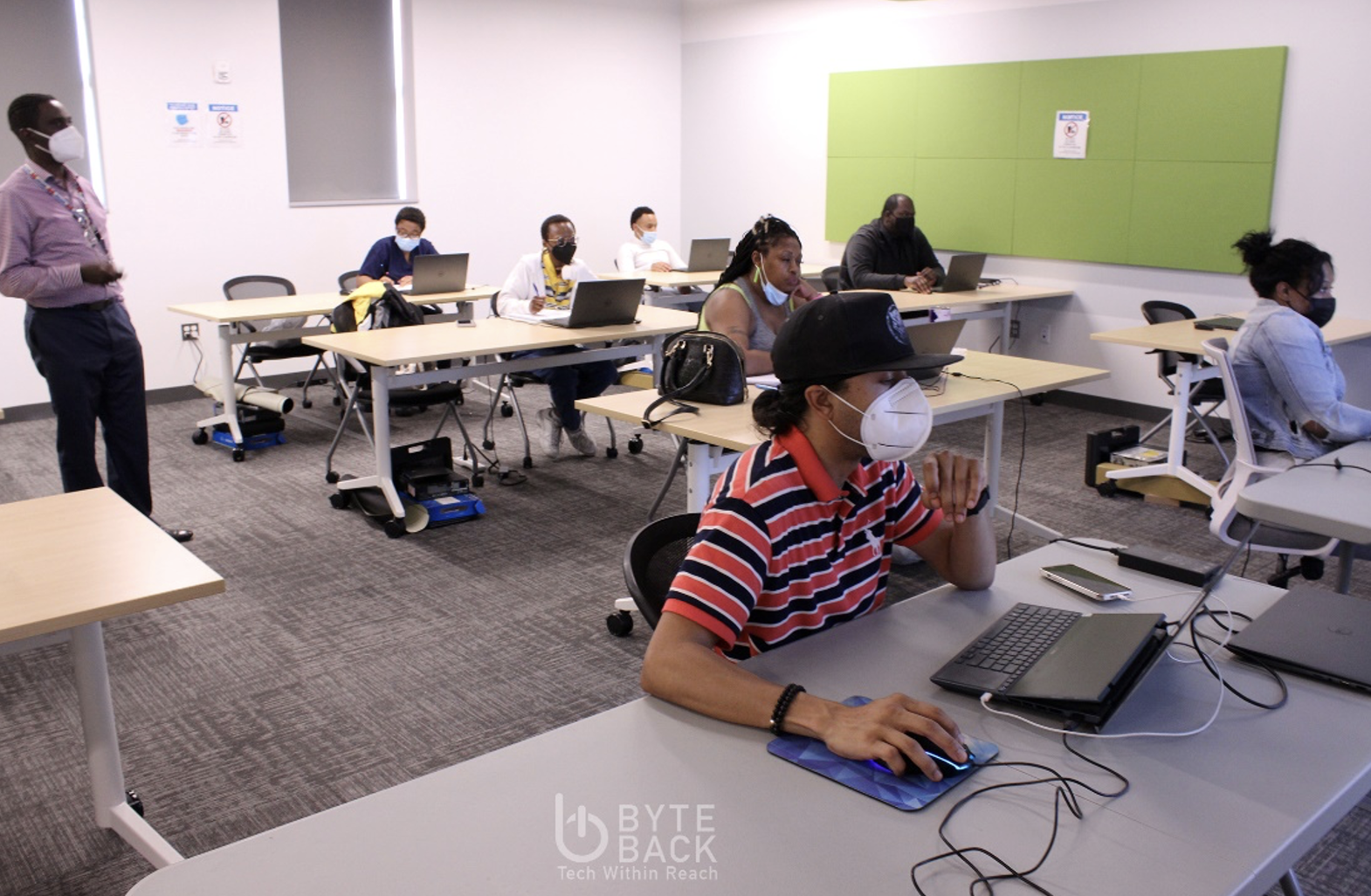 Byte Back scholars are seated in a training, working on individual laptops. An instructor stands to the side of the classroom.